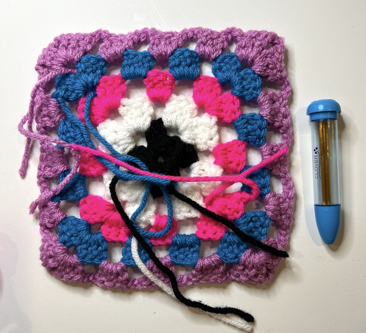 Granny square with all of the yarn ends hanging off from the wrong side with the needles in their case