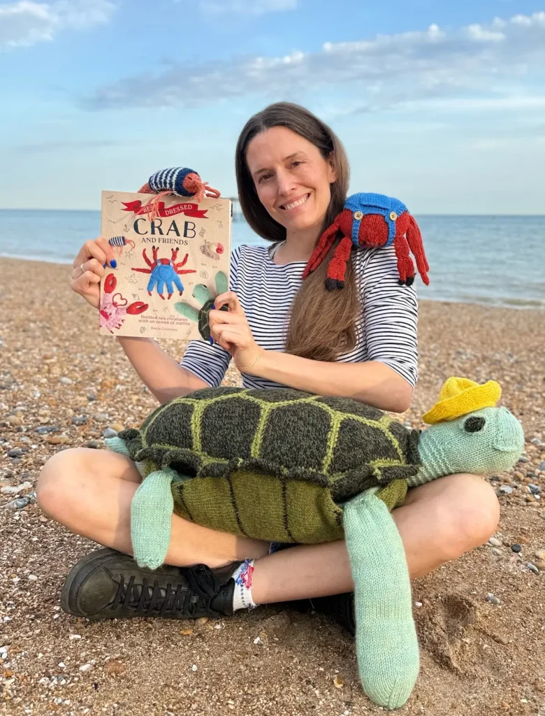 Karla Courtney on the beach holding her book Crab and Friends with a knitted turtle on her lap
