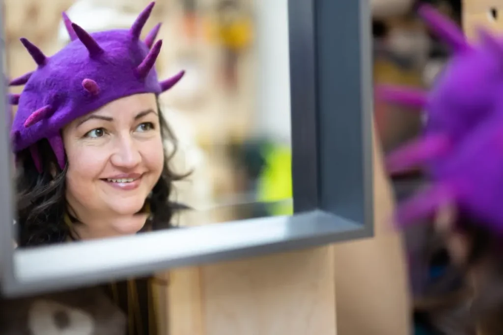 Niki Collier in a purple hat looking in a mirror and smiling