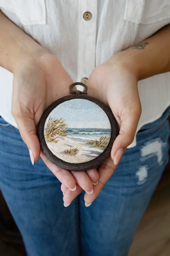 Cassandra Dias holding a small embroidery art of sand dunes and ocean